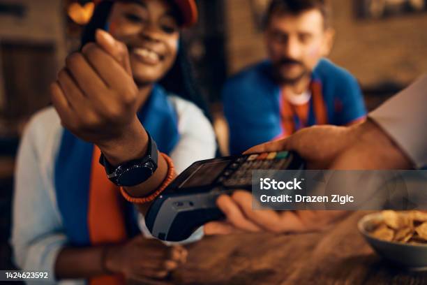 Close Up Of Black Sports Fan Using Her Smart Watch While Paying Contactless In Pub Stock Photo - Download Image Now