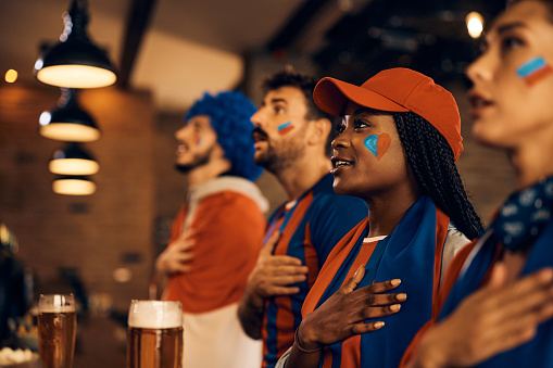 African American soccer fan and her friends with hand on heart singing national anthem while watching sports championship in bar.