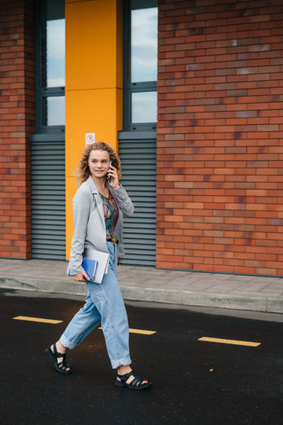 istock Young caucasian college student holding laptop computer, talking on cell phone while walking on street outside office. Looking aside talking by phone. A full-length portrait. 1424629124