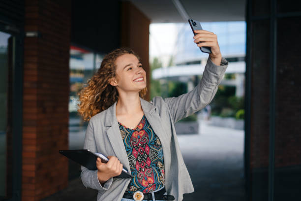 Beautiful happy young caucasian woman student taking a selfie photo with smart phone holding laptop outdoors in downtown city center. People, millennial generation and technology concept. Selfie mania! stock photo