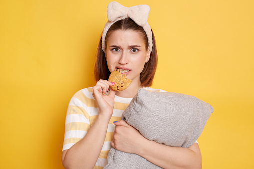 Portrait of young adult dark haired woman biting cookie eating sweets isolated on yellow background, being in bad mood, looking at camera with sad expression.