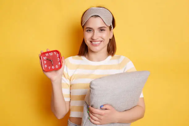 Indoor shot of impressed dark haired woman stares at camera prepares for sleep holds soft pillow and red alarm clock dressed in sleepmask on forehead isolated over yellow background.