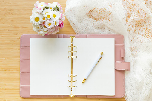 Flat lay, top view romantic setup of a pastel pink planner, flower bouquet and stationery on a wooden desk. Planning, writing, blogging.
