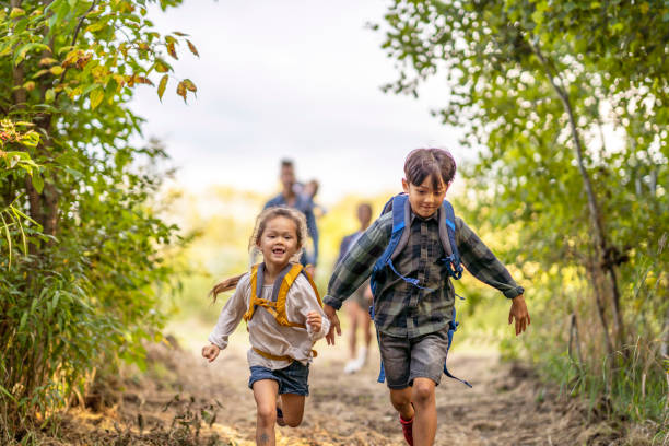 A small family of five hike together on a warm summer day.  They are each dressed casually and have backpacks on with their supplies for the day.  The father is carrying the youngest child in an carrier on his back.