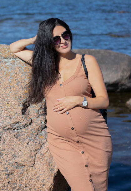 Heavily pregnant beautiful brunette woman with black sunglasses and beige tight dress leaning against a big boulder on the beach with a blue sea behind her on a sunny summer day stock photo