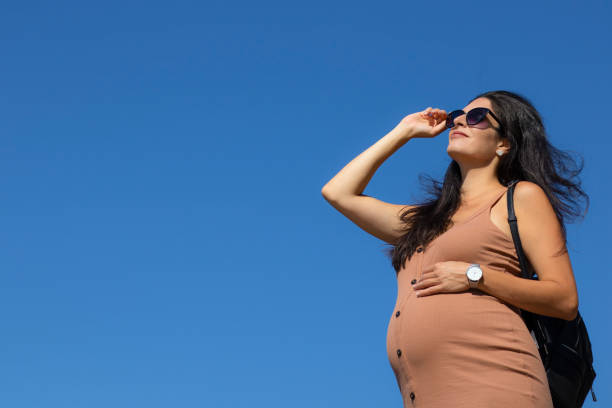 Nine months pregnant brunette woman in a beige dress, black sunglasses and a backpack touching her tummy and looking upwards into the distance on the clear blue sky background stock photo