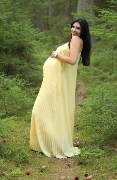 Heavily pregnant attractive and smiling brunette woman in her 30s in a long yellow maxi dress standing on a path in the woods and posing in the forest full of spruces and evergreen trees stock photo