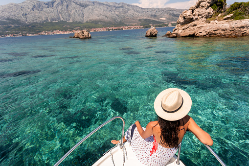 A beautiful, blonde woman with a hat sits on a boat and enjoys the calm sea during her summer holidays