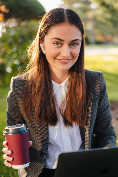 portrait of young female holding a cup of coffee looking at camera while working on laptop, sitting on grass in park on sunny day outdoors. relax in sunshine green city park outdoors on nature. urban leisure concep - surfing wireless vertical outdoors lifestyles imagens e fotografias de stock