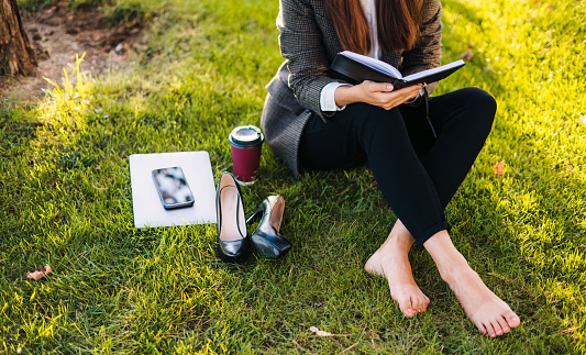 Cropped shot of young creative businesswoman checking plans for week in diary sitting on grassy lawn in the park. Laptop, coffee cup, pencil, mobile phone. The girl works, studies, learning, keeps a diary.
