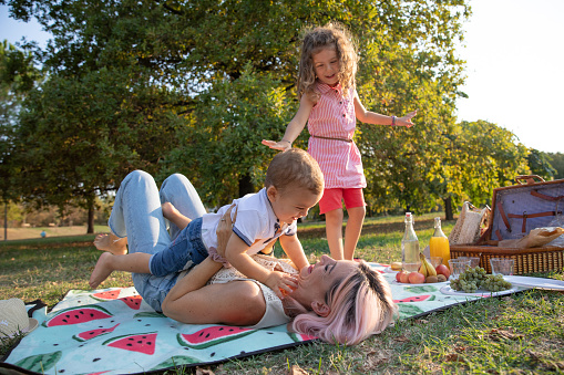Mother at a picnic plays with her children, lifts her son while her daughter watches, fruits and vegan food at the picnic.