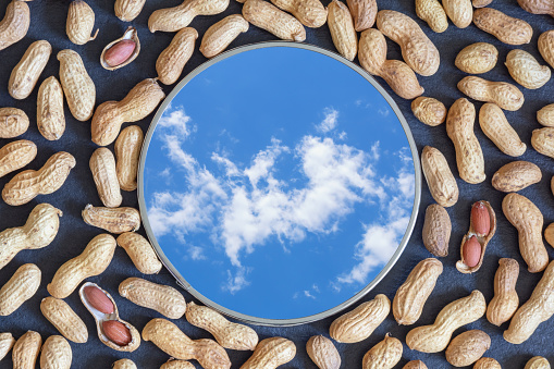 Peanuts and mirror on dark table. Mirror reflects blue sky and white clouds. Flat lay
