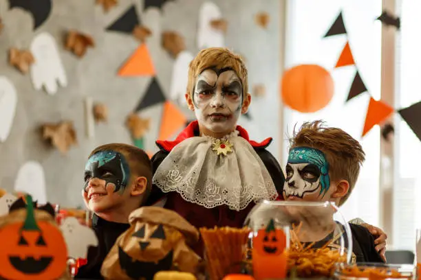 Front view of cute boy, in Dracula costume and face paint, embracing his two little friends, also wearing costumes and face paint and looking at camera while having fun on a Halloween party.