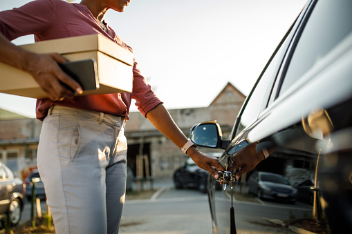 Midsection of unrecognizable woman standing by her car, holding a package and opening a door by the handle.