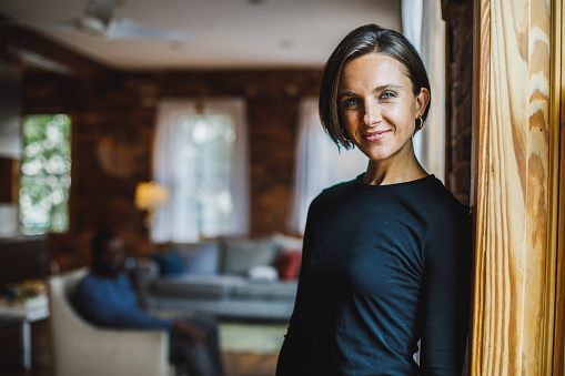 Portrait of happy mid adult woman, with piercing blue eyes, leaning against the brick wall, looking at camera and smiling with confidence. Her partner hanging out in the background.