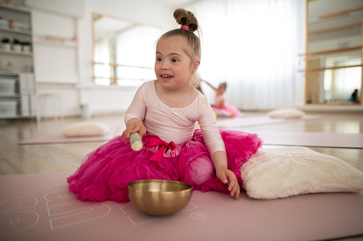 Little girl with down syndrom sitting on the floor in a ballet dancing studio and making sound on a tibetian singing bowl.