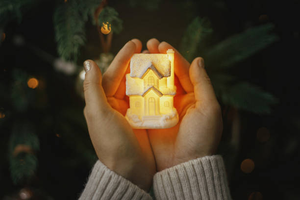 Little glowing house in hands on background of illuminated christmas tree lights bokeh. Magical holiday time. Cozy home. Atmospheric  image. Family togetherness and safety concept. Merry Christmas! stock photo