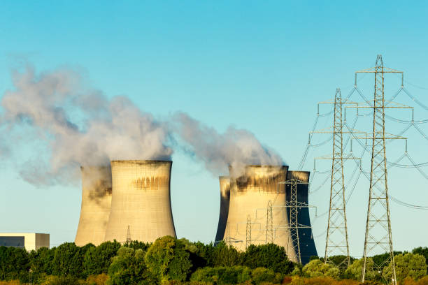 Electricity towers or Pylons linking into a Power Station with water vapour plumes rising from the large cooling towers, against a clean blue sky. stock photo
