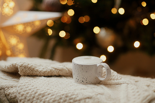 Warm cup of coffee on background of christmas tree with lights. Cozy home. Atmospheric winter hygge. Stylish mug on knitted sweater against golden illumination bokeh. Christmas banner