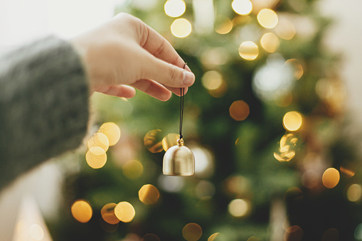 Hand holding golden bell on background of illuminated christmas tree with golden lights. Woman decorating christmas tree and holding glitter ornament in hand in festive room. Atmospheric time