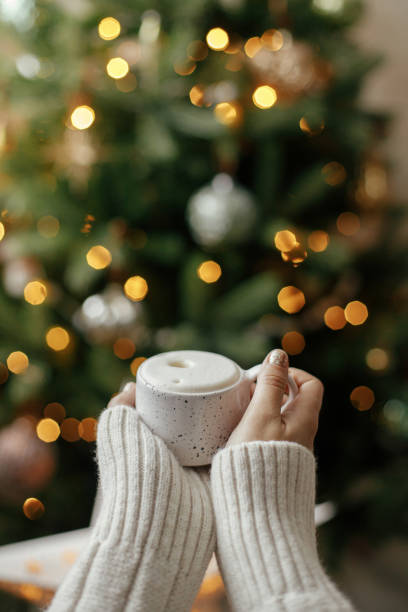Hands holding warm cup of coffee on background of christmas tree with lights. Cozy home, atmospheric winter hygge. Woman hands in warm sweater holding  stylish mug at illumination bokeh stock photo