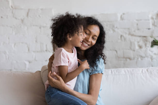 African mom sit on sofa hugging cheerful little daughter