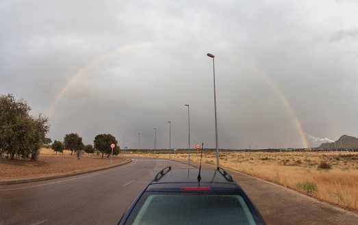 Car at roundabout with rainbow rising over horizon. Industrial area