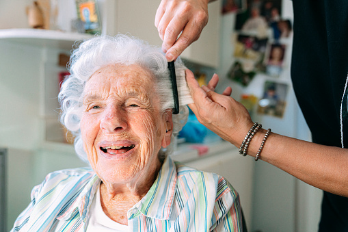 Wide Angle Shot of a Cheerful, Cute Elderly Senior Caucasian Woman at Home having Her Hair Combed and Teased by a Professional In-Home Caregiver Hairdresser During Her Weekly Shampoo Set Hair Appointment