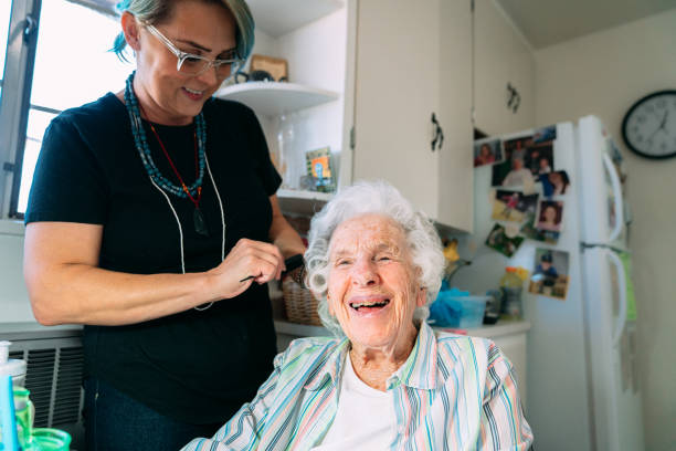 Wide Angle Shot of a Cheerful, Cute Elderly Senior Caucasian Woman at Home having Her Hair Combed by a Professional In-Home Caregiver Stylist stock photo