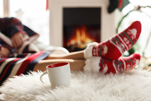 Selective focus shot of a cozy fireplace, unrecognizable young woman wearing warm Christmas themed socks and enjoying a hot beverage.