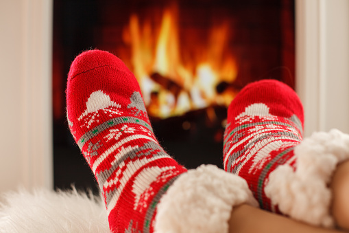 Close up shot of unrecognizable woman keeping her feet warm by the fireplace while waring fuzzy Christmassy socks.
