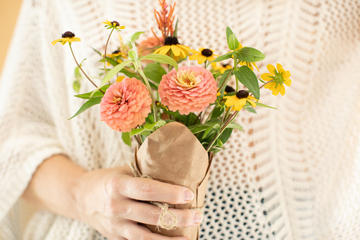 Woman's hand holding a small jar covered with brown paper full of fresh cut yellow and pink flowers.