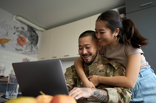 Joyful asian woman in kitchen room hugging smiling military asian man from behind in front of laptop