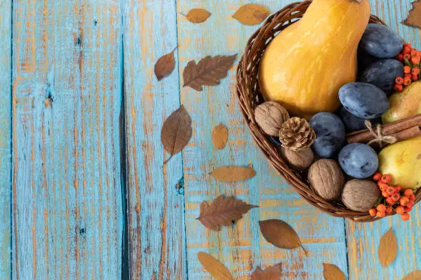 Autumn fruit (butternut squash, plum, pear, walnut, cinnamon) in a wicker basket on a rustic wooden background with dry leaves. Copy space. Top table view. Thanksgiving day, fall harvesting concept.