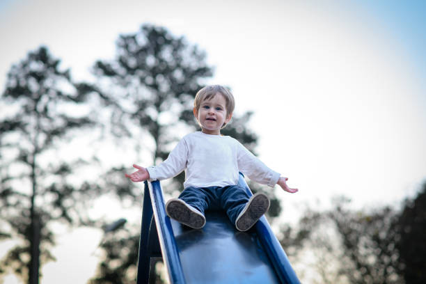 Toddler boy looking at camera with open arms getting ready to slide down slide at playground at sunset