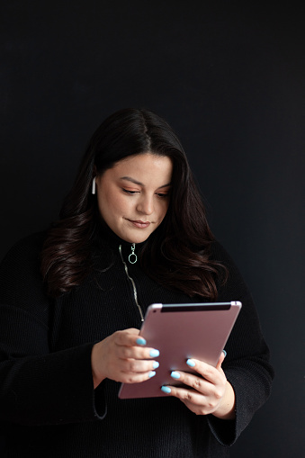 Portrait of a young beautiful plus size woman wearing wireless headphones, holding and using her digital tablet. Voluptuous young businesswoman posing in front of a black wall.