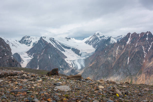 istock Dramatic landscape with high snowy mountain range with long glacier tongue and pointed peak in sunlight under cloudy sky. Atmospheric view from stone hill to large snow mountains at changeable weather 1424745042