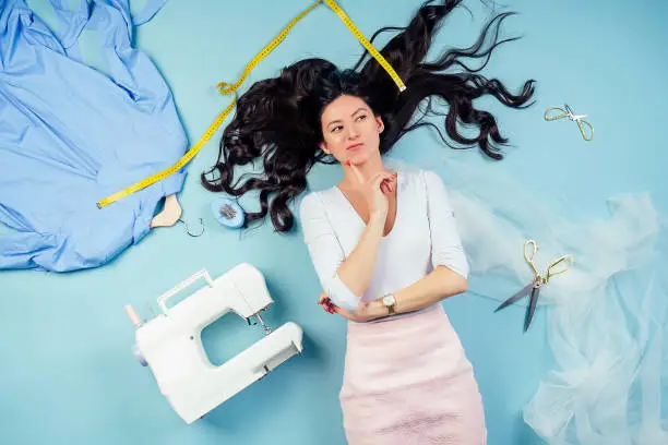 attractive brunette woman seamstress tailor ( dressmaker) dreams and thinks about a new collection of clothes on the floor with sewing machine and measuring tape on a blue background in the studio.