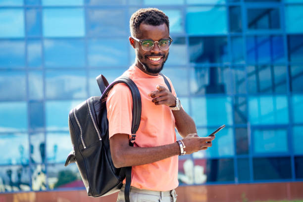 guapo afroamericanos de moda nerd masculino con gafas en ropa elegante camiseta colorida con una mochila en los hombros de pie contra el fondo de las ventanas azules de la universidad - male education college student nerd fotografías e imágenes de stock