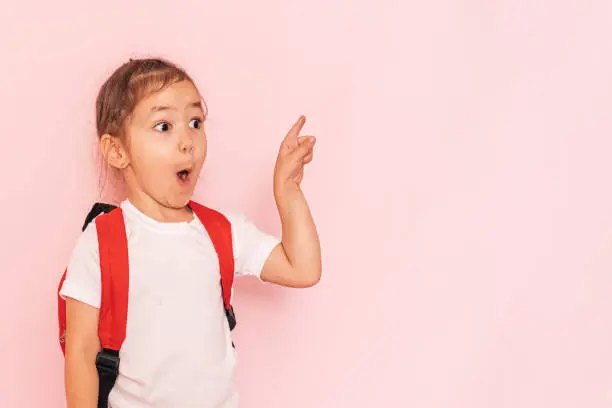 Photo of Surprised little girl with a red briefcase points her finger against a pink background