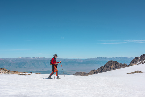Tourist in red walks on snow mountain near abyss edge on high altitude under blue sky in sunny day. Man with camera on snowy mountain near precipice edge with view to large mountain range in away.