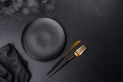An empty black plate on a dark concrete background with spices, herbs and cutlery. Home dinner cooking