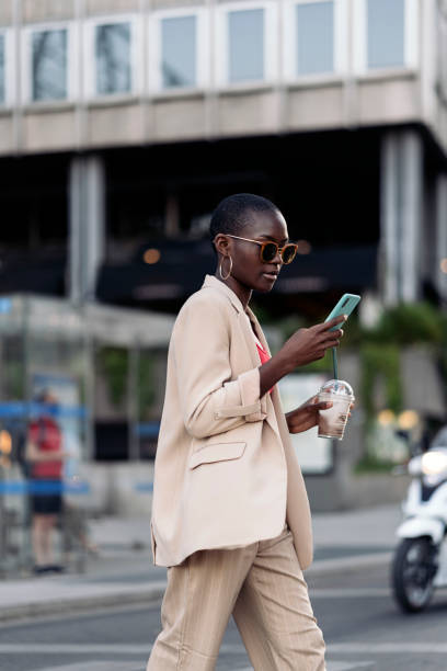 elegante giovane donna afroamericana che cammina per strada e usa il telefono mentre tiene in mano un frappè - blueberry smoothie milk shake drink foto e immagini stock