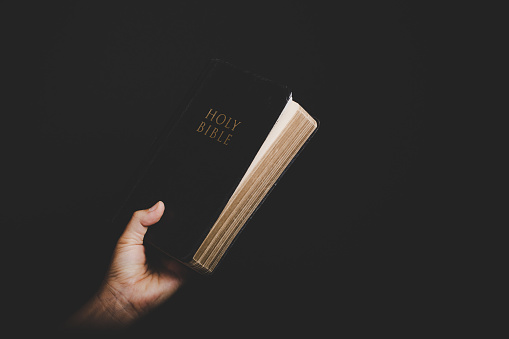 High contrast shot of a minister holding an open Bible and pointing at you.