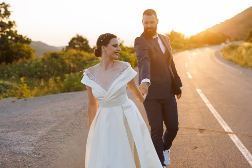 Romantic just married young couple holding hands and  walking on empty road in nature at sunset