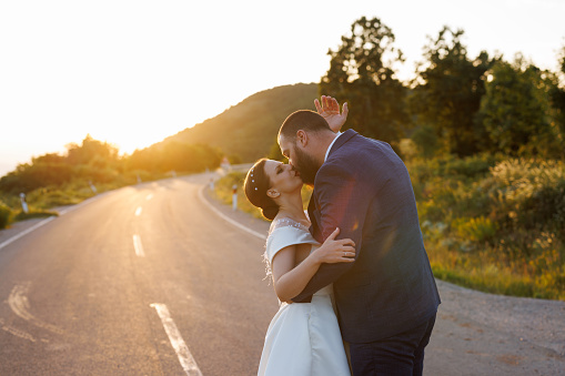 Romantic newlyweds kissing on empty road in nature at sunset