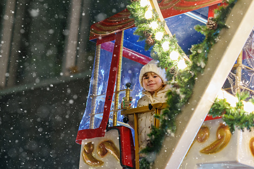 Little preschool girl riding on ferris wheel carousel horse at Christmas funfair or market, outdoors. Happy toddler child having fun on traditional family xmas market in Germany