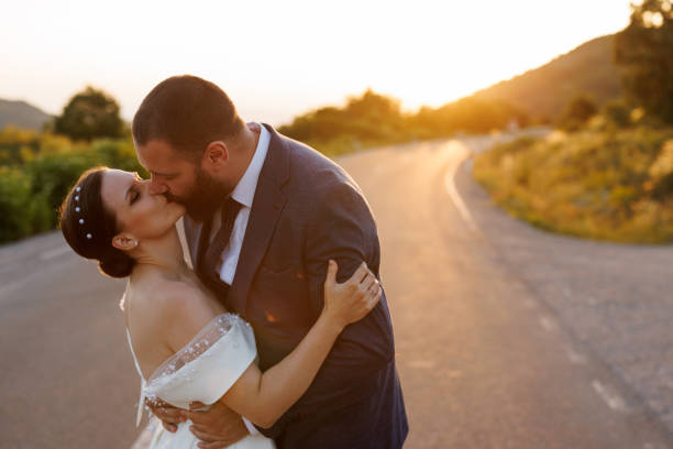 Romantic newlyweds kissing on empty road in nature at sunset