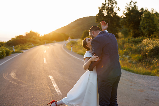 Romantic just married young couple dancing and kissing on empty road in nature at sunset