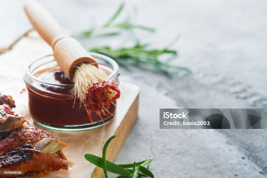Hot and spicy barbecue sauce in jar bowl with basting brush on old stone gray background. Flat lay composition. American food concept. Top view. Space for text Barbeque Sauce Stock Photo